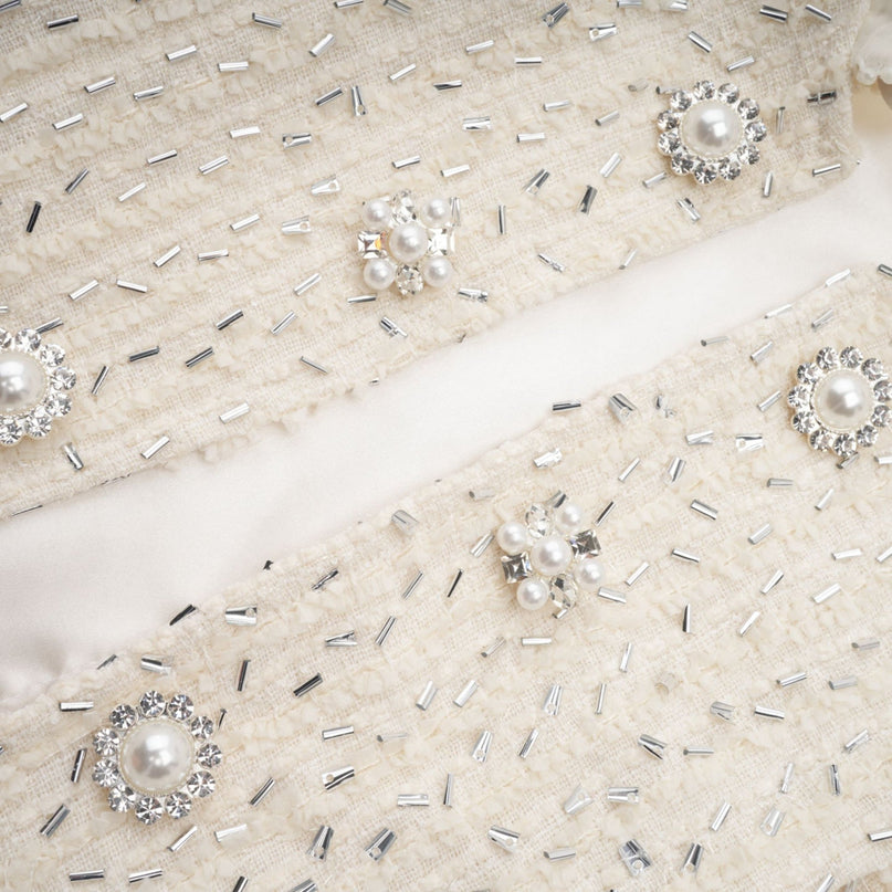 A white cropped jacket with beaded details and ruffled trim worn by a model against a plain background