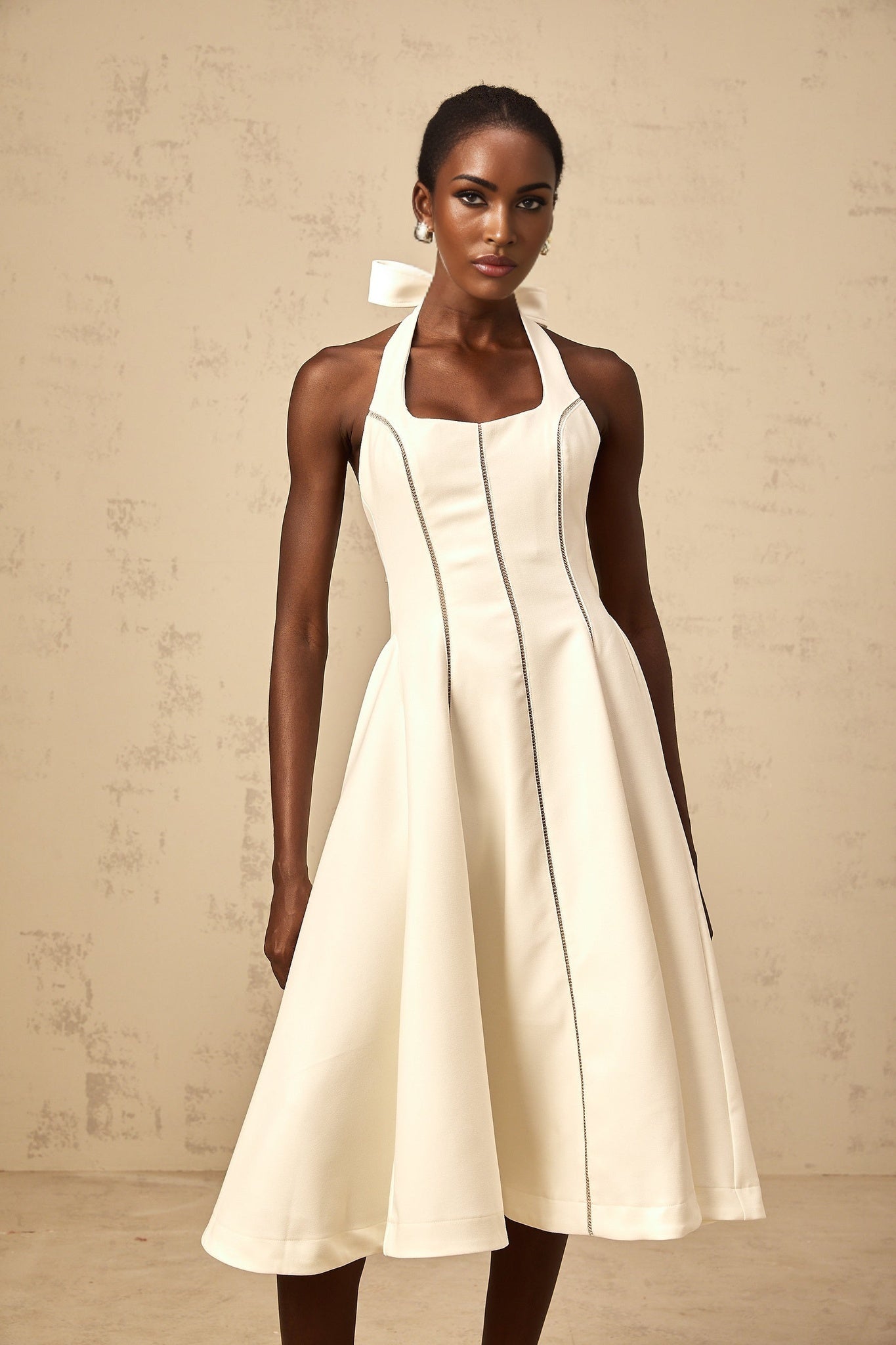 A white halterneck midi dress with a bow detail worn by a model standing against a plain background