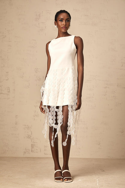 A white midi dress with a disc design pattern and fringed details worn by a model standing against a plain background