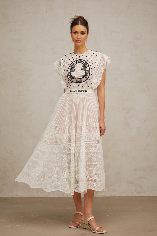 A white midi dress with a pleated skirt featuring jacquard floral lace pattern named Phoebe worn by a model standing against a plain background