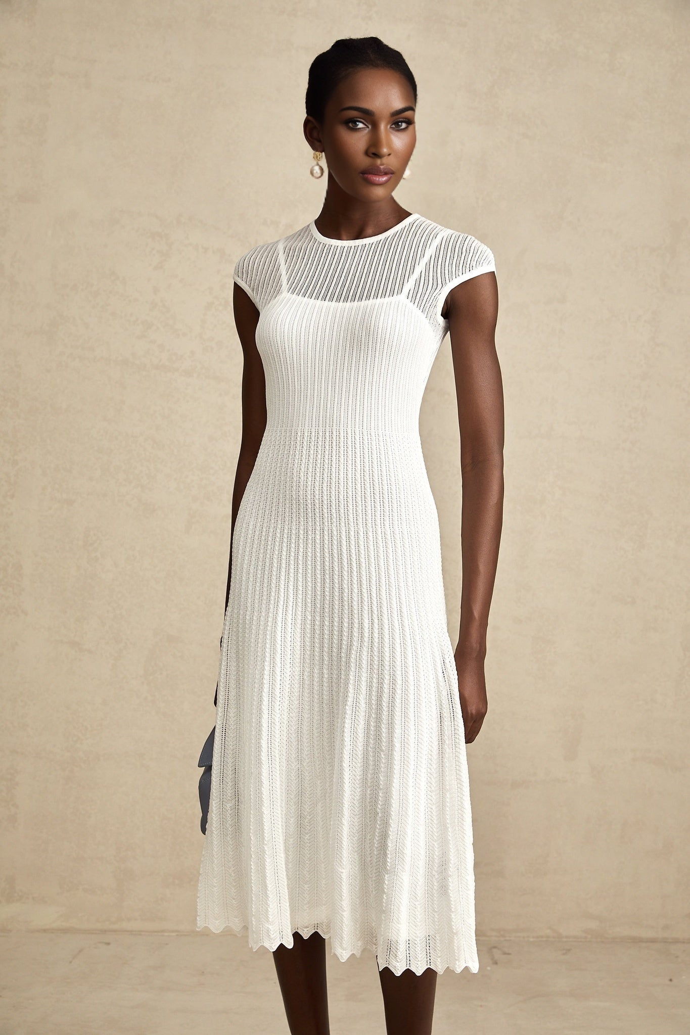 A white midi dress with a scalloped hem and semi sheer fabric worn by a model standing against a plain background