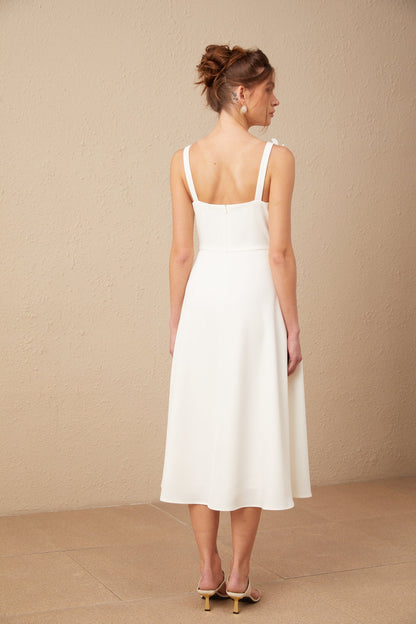 A white midi dress with a square neckline featuring faux flower details worn by a model against a plain background