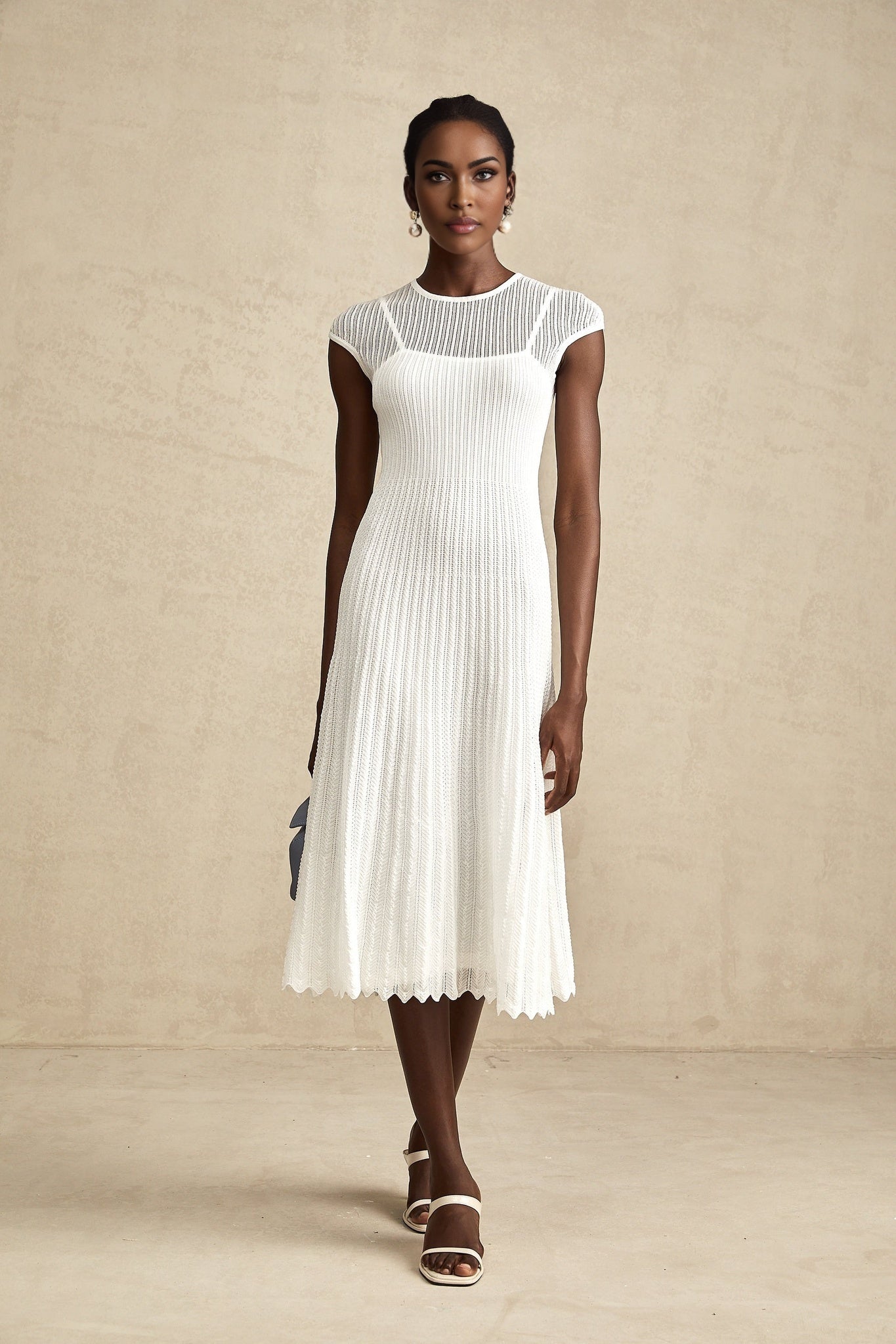 A white midi dress with scalloped hem and semi sheer fabric worn by a model standing in a neutral background