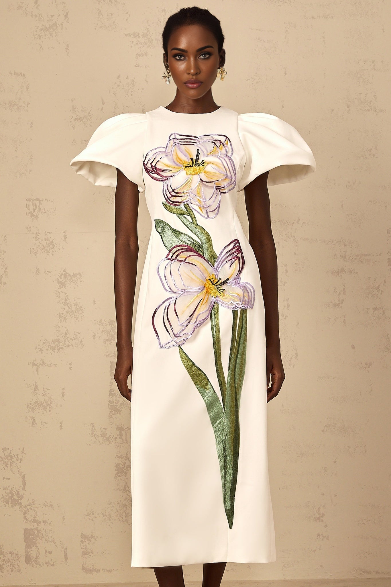 A white midi dress with sequin floral embroidery worn by a woman standing against a plain background