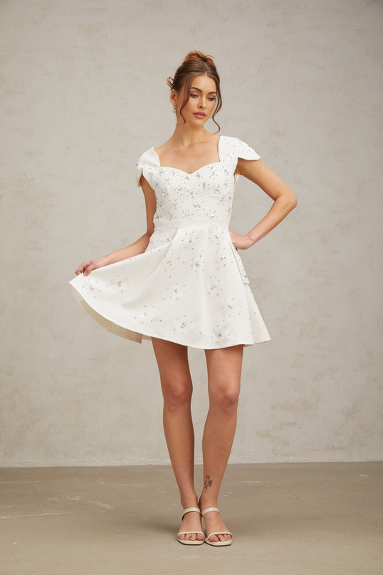 A white mini dress with a flared skirt featuring embellishments and named Bonnie worn by a model against a plain background