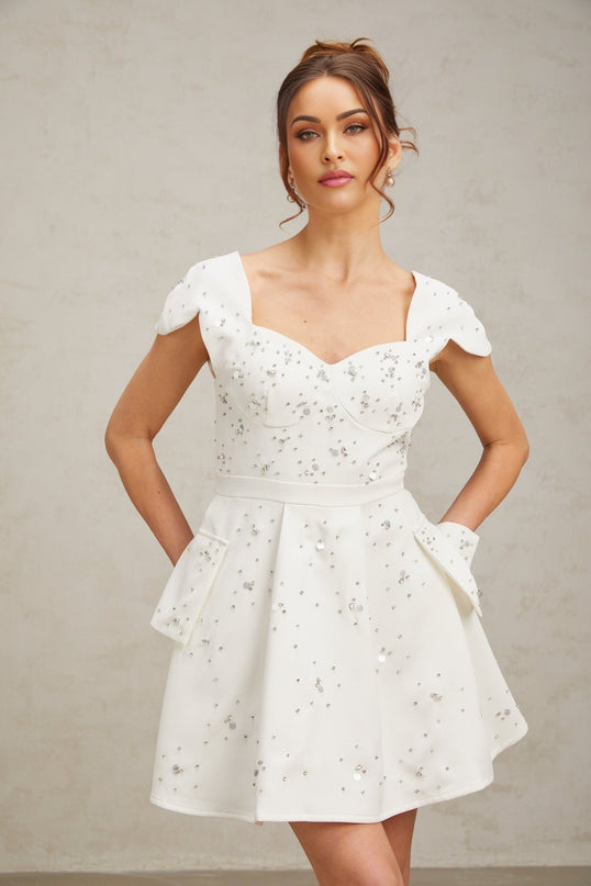 A white mini dress with a flared skirt featuring embellishments named Bonnie worn by a model against a plain background