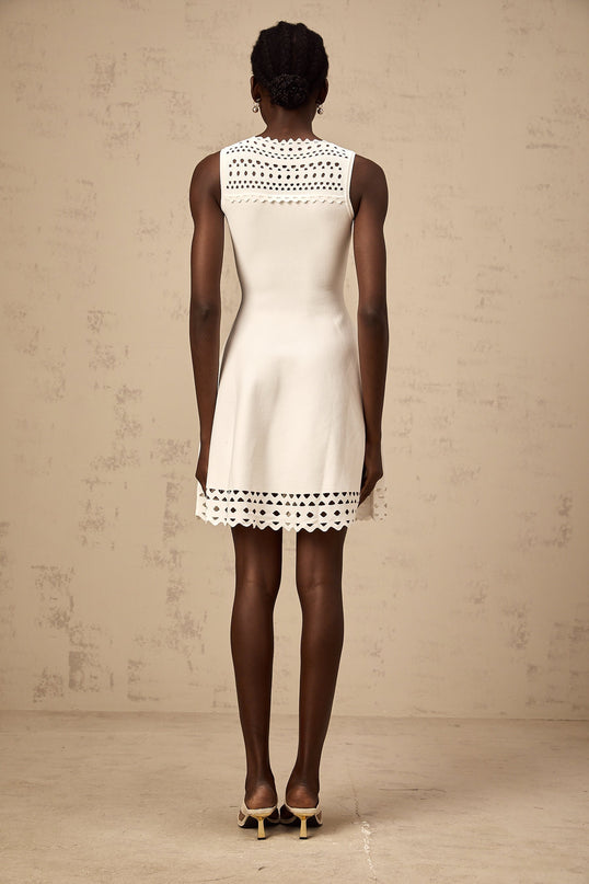 A white sleeveless mini dress with cutout details and a knitted texture worn by a model standing against a plain background