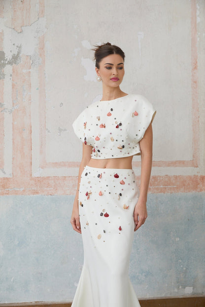 A white twopiece outfit featuring a top and skirt adorned with decorative shell embellishments both pieces matching to create a coordinated set