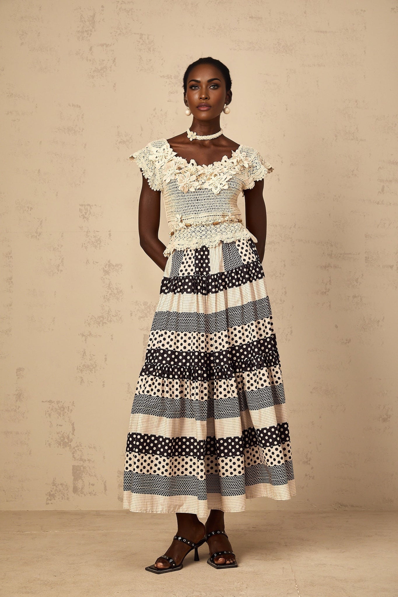 A woman wearing a beige crochet knit top with delicate three dimensional floral petal details and intricate beadwork along the neckline and sleeves