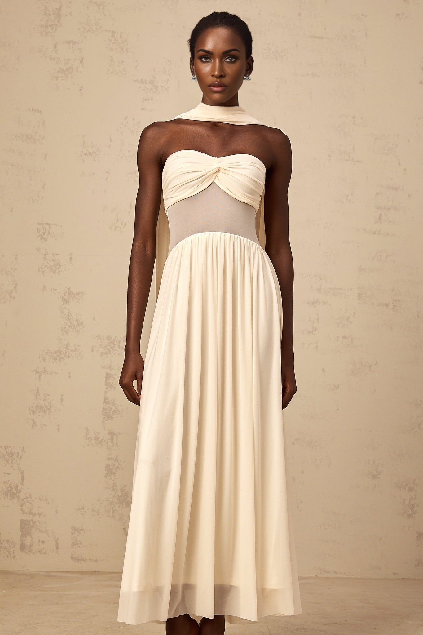 A woman wearing a beige midi dress with a scarflike design and mesh detailing standing against a neutral background
