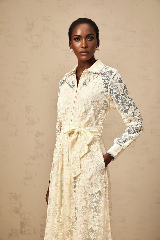 A woman wearing a beige midi length shirt dress with delicate guipure lace details standing in a neutral background