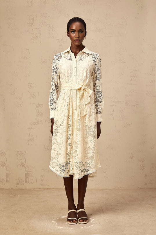 A woman wearing a beige midi length shirt dress with intricate guipure lace detailing standing against a plain background