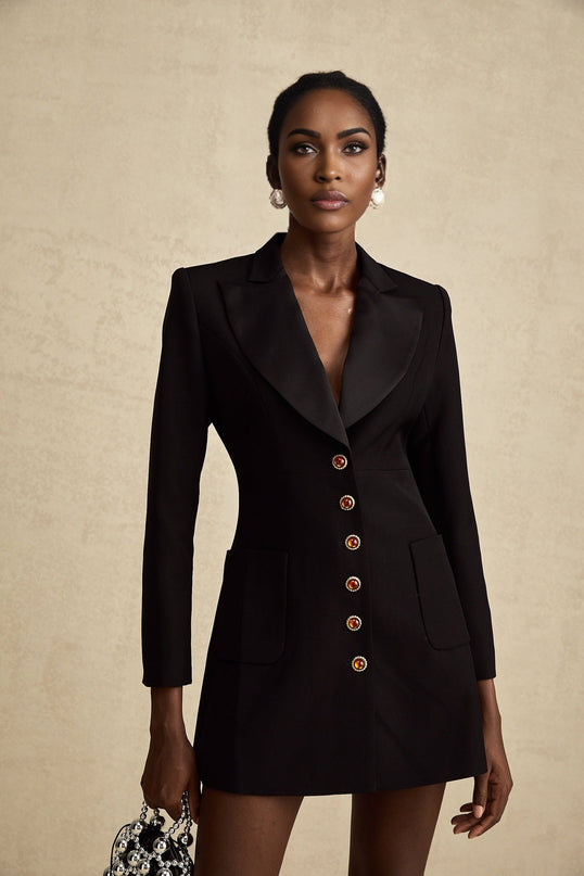 A woman wearing a black blazer dress with a singlebreasted design standing against a plain background