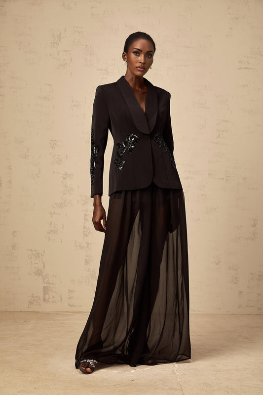 A woman wearing a black blazer with sheer tulle panels and matching trousers both embellished with sparkling details standing against a plain background