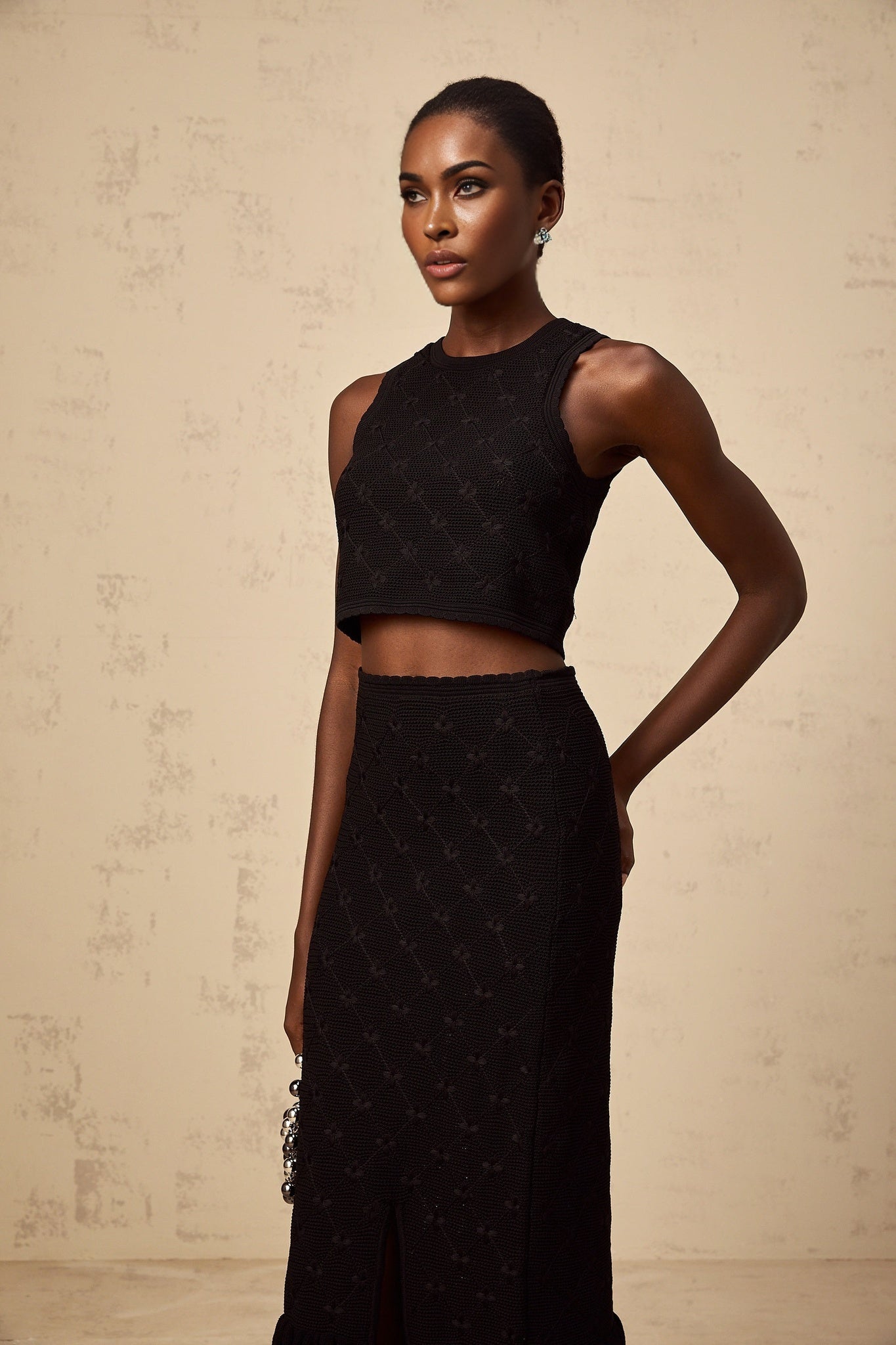 A woman wearing a black crochet knit matching set consisting of a sleeveless top and a short skirt standing against a plain background
