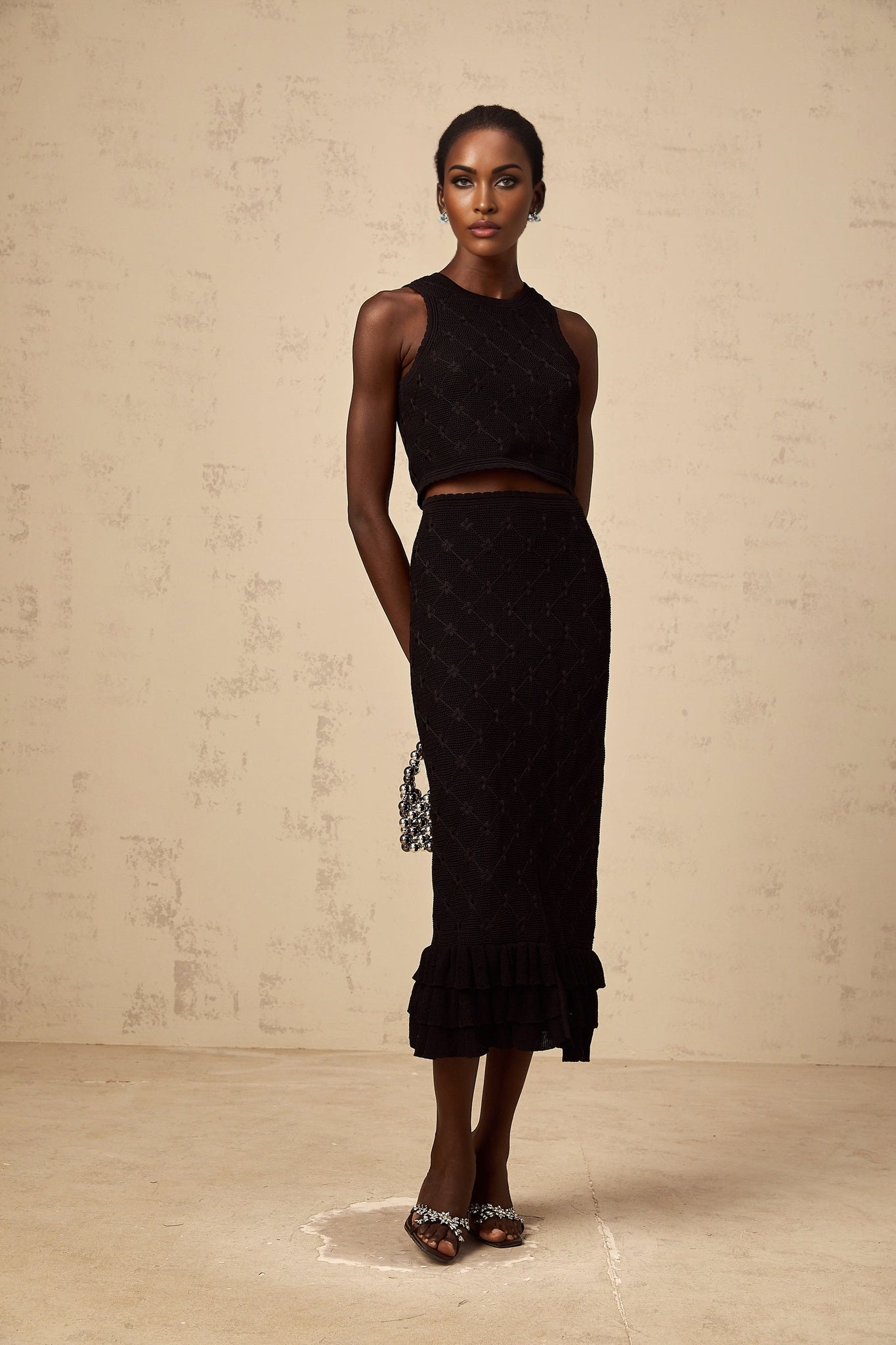 A woman wearing a black crochet knit matching set consisting of a sleeveless top and a short skirt standing against a plain background