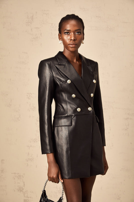 A woman wearing a black leather blazer mini dress with long sleeves and a tailored fit standing against a plain background