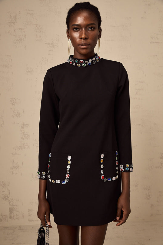 A woman wearing a black mini dress with long sleeves adorned with crystal embellishments standing in an elegant pose