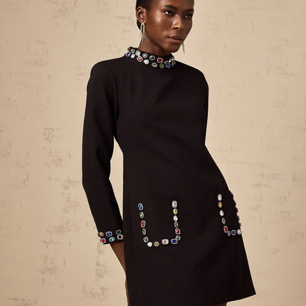 A woman wearing a black mini dress with long sleeves and crystal embellishments standing in a neutral pose