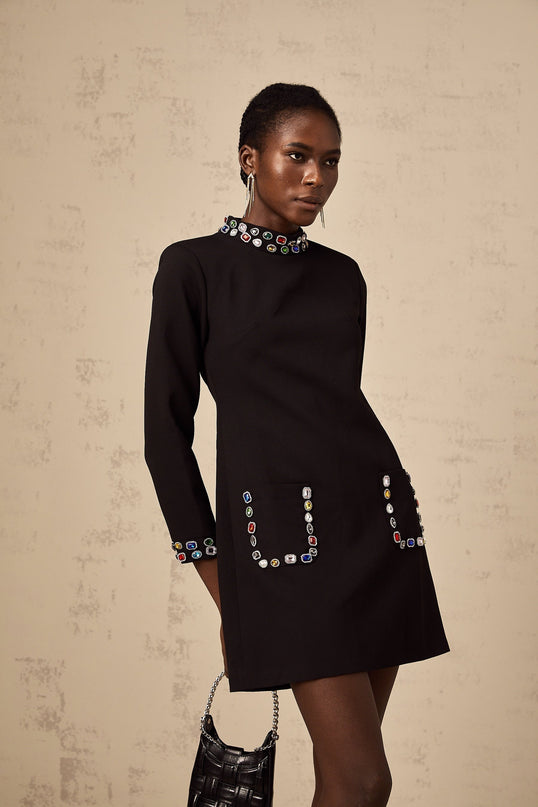 A woman wearing a black mini dress with long sleeves and crystal embellishments standing in a neutral pose