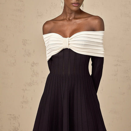 A woman wearing a black offshoulder knit mini dress with a bow detail standing in a neutral setting