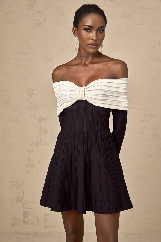 A woman wearing a black offshoulder knit mini dress with a bow detail standing in a neutral setting