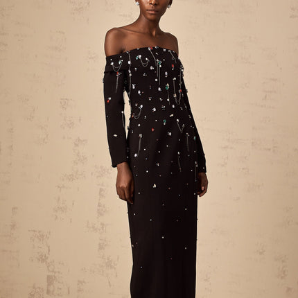 A woman wearing a black offshoulder maxi dress with crystal embellishments standing elegantly against a plain background