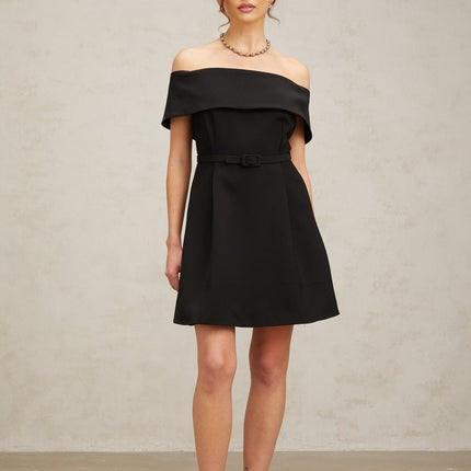 A woman wearing a black offshoulder mini dress with a belt cinching the waist standing against a plain background