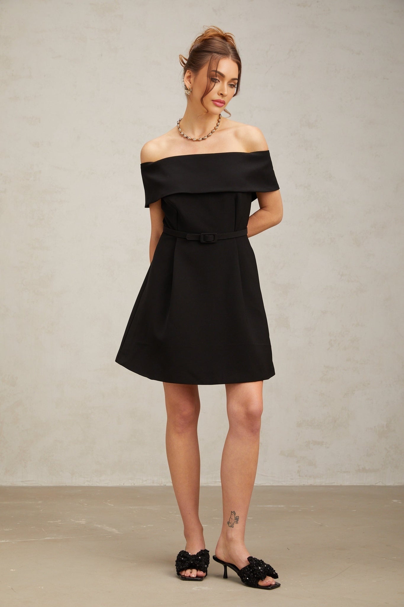 A woman wearing a black offshoulder mini dress with a belt cinching the waist standing against a plain background
