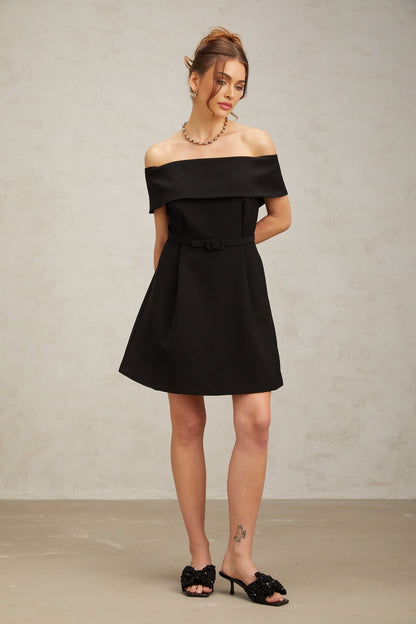 A woman wearing a black offshoulder mini dress with a belt cinching the waist standing against a plain background