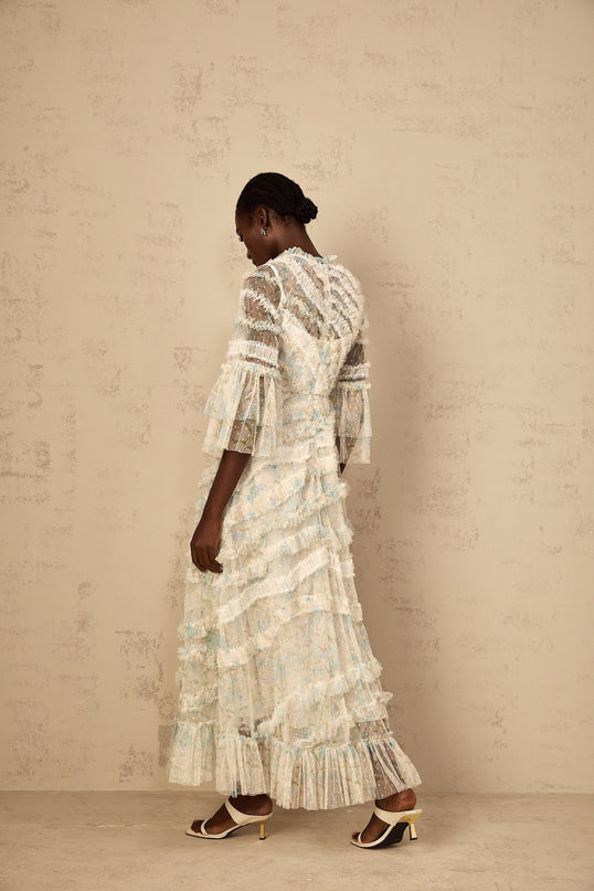 A woman wearing a blue maxi dress with a floral print and ruffled tulle details standing against a plain background