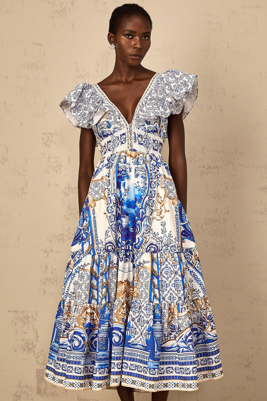A woman wearing a blue midi dress with a baroque print pattern and tiered design standing in a neutral background
