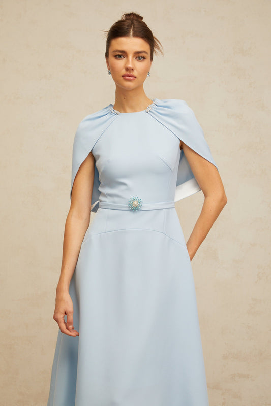 A woman wearing a blue midi dress with shawl panels and embellishments standing in a neutral background