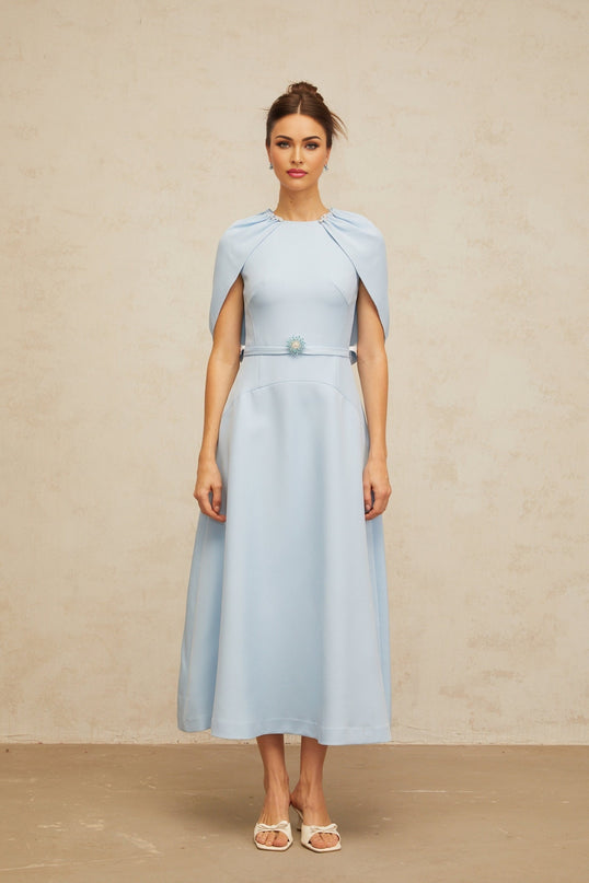 A woman wearing a blue midi dress with shawllike panels and embellishments posing against a plain background