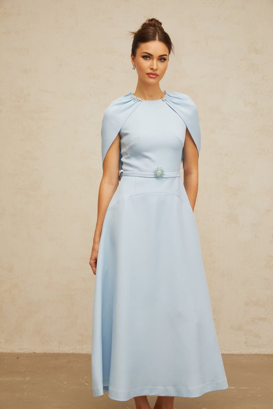 A woman wearing a blue midi dress with shawllike panels and embellishments standing in a relaxed pose