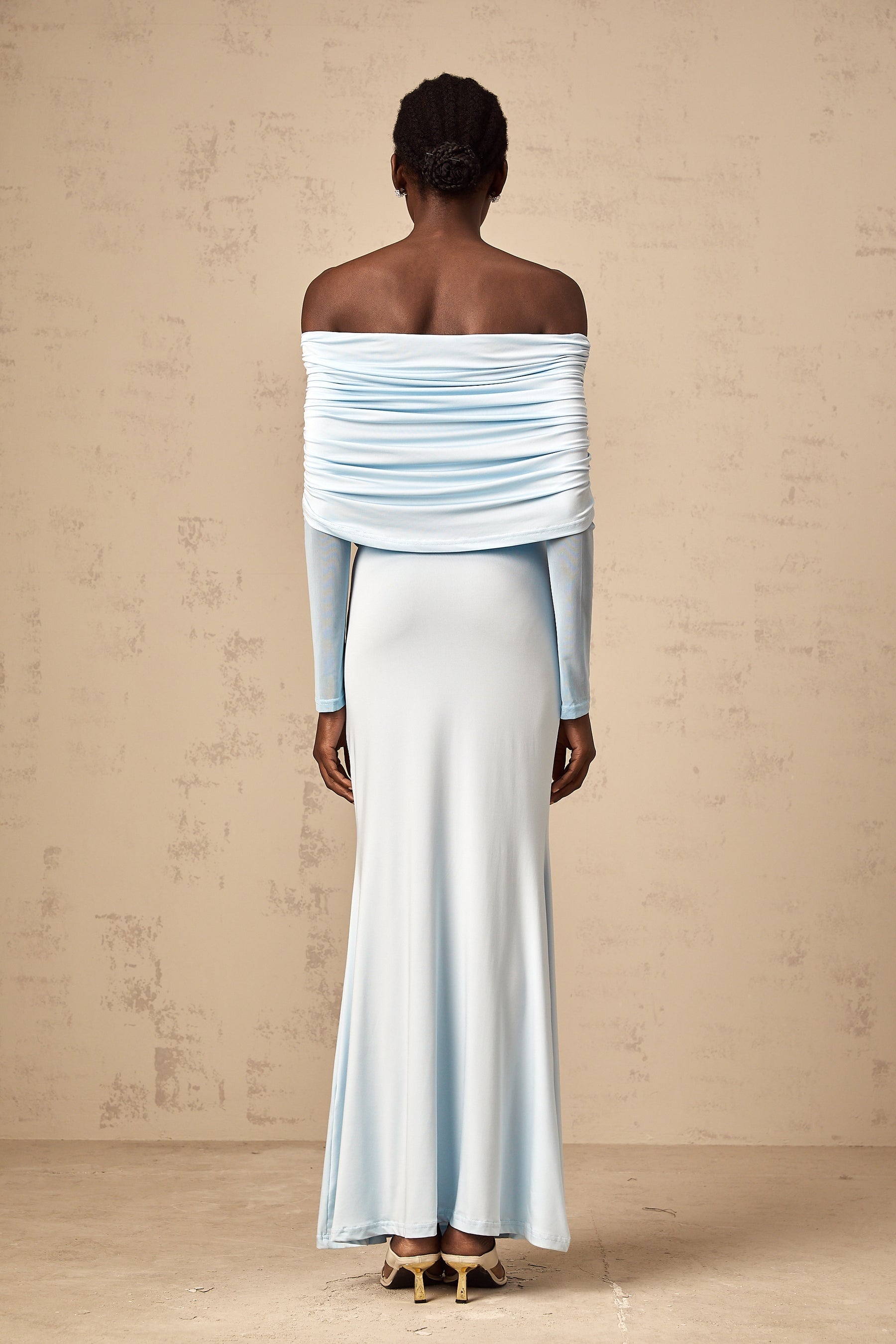 A woman wearing a blue offshoulder maxi dress with ruched detailing standing against a plain background