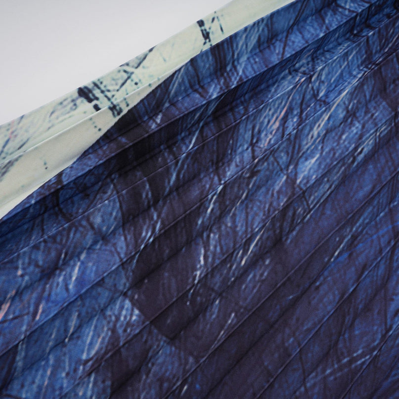 A woman wearing a blue patterned cape and matching skirt set The cape is creased and flowy while the skirt is fitted The pattern features abstract geometric shapes in various shades of blue The outfit is styled on a model against a plain background