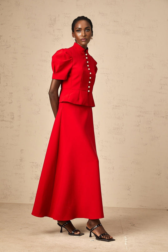 A woman wearing a bright red matching set consisting of a pleated blouse and skirt The blouse has long sleeves and a high neck while the skirt falls to mid length Both pieces feature pleated detailing for a coordinated look