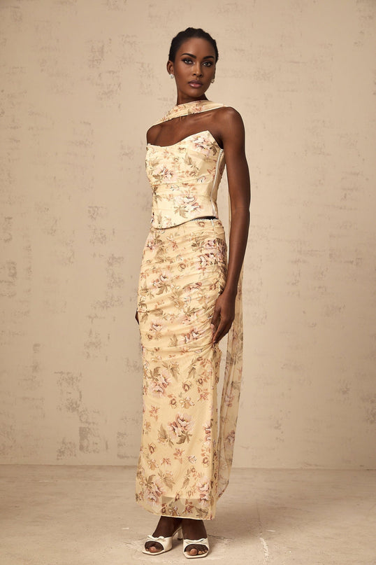 A woman wearing a bright yellow floral print matching set consisting of a fitted top and skirt standing against a plain background