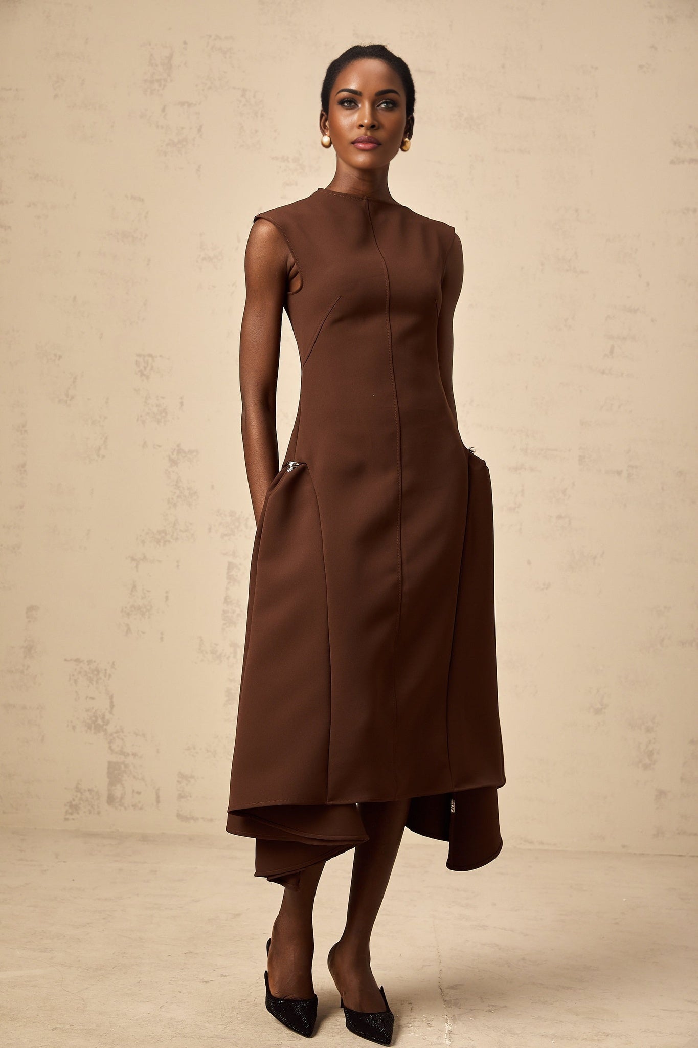A woman wearing a brown asymmetric midi dress with a flowy skirt and one shoulder design