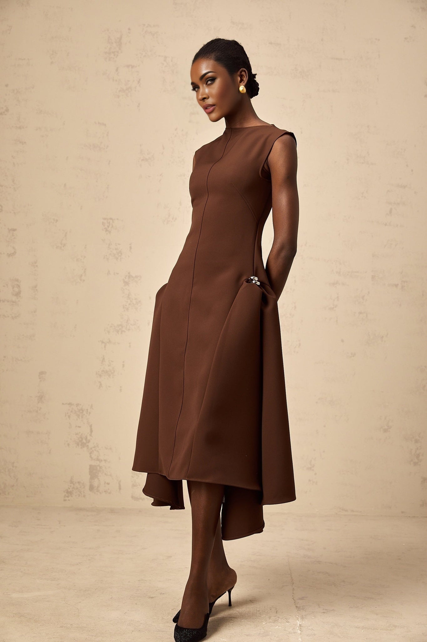 A woman wearing a brown asymmetrical midi dress with a draped neckline and flowing skirt standing against a plain background