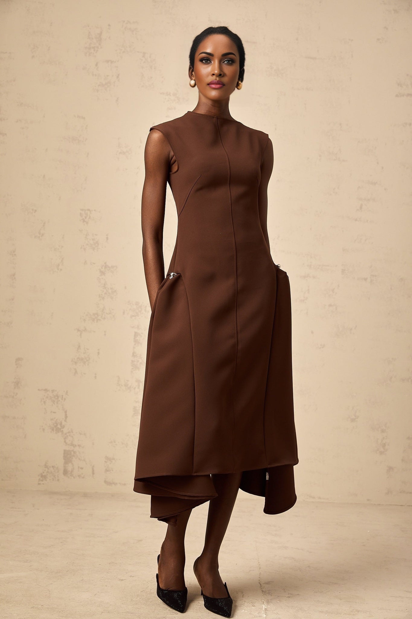 A woman wearing a brown asymmetrical midi dress with a flowing design standing against a plain background