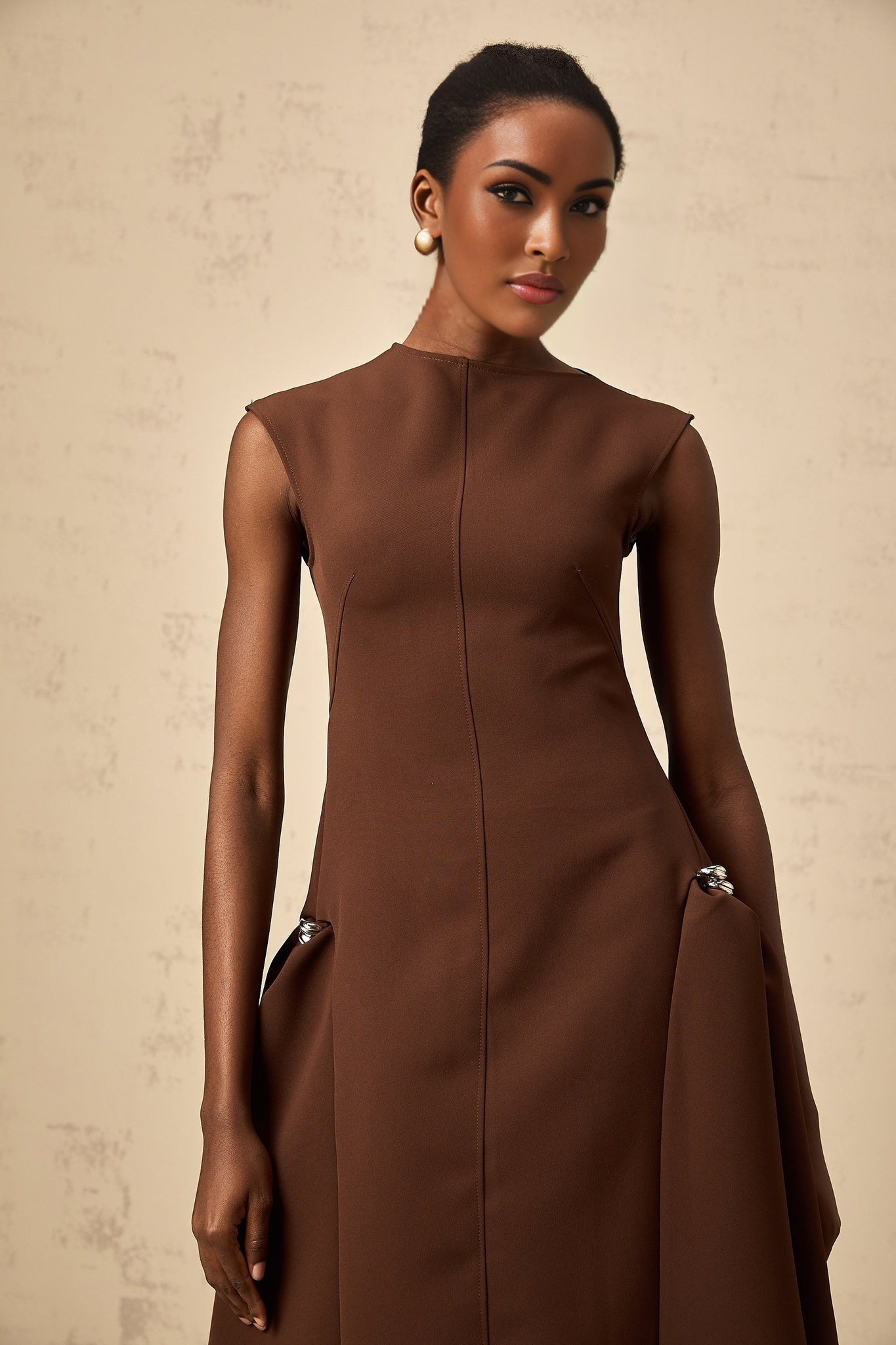 A woman wearing a brown asymmetrical midi dress with a flowing skirt and fitted bodice standing against a plain background