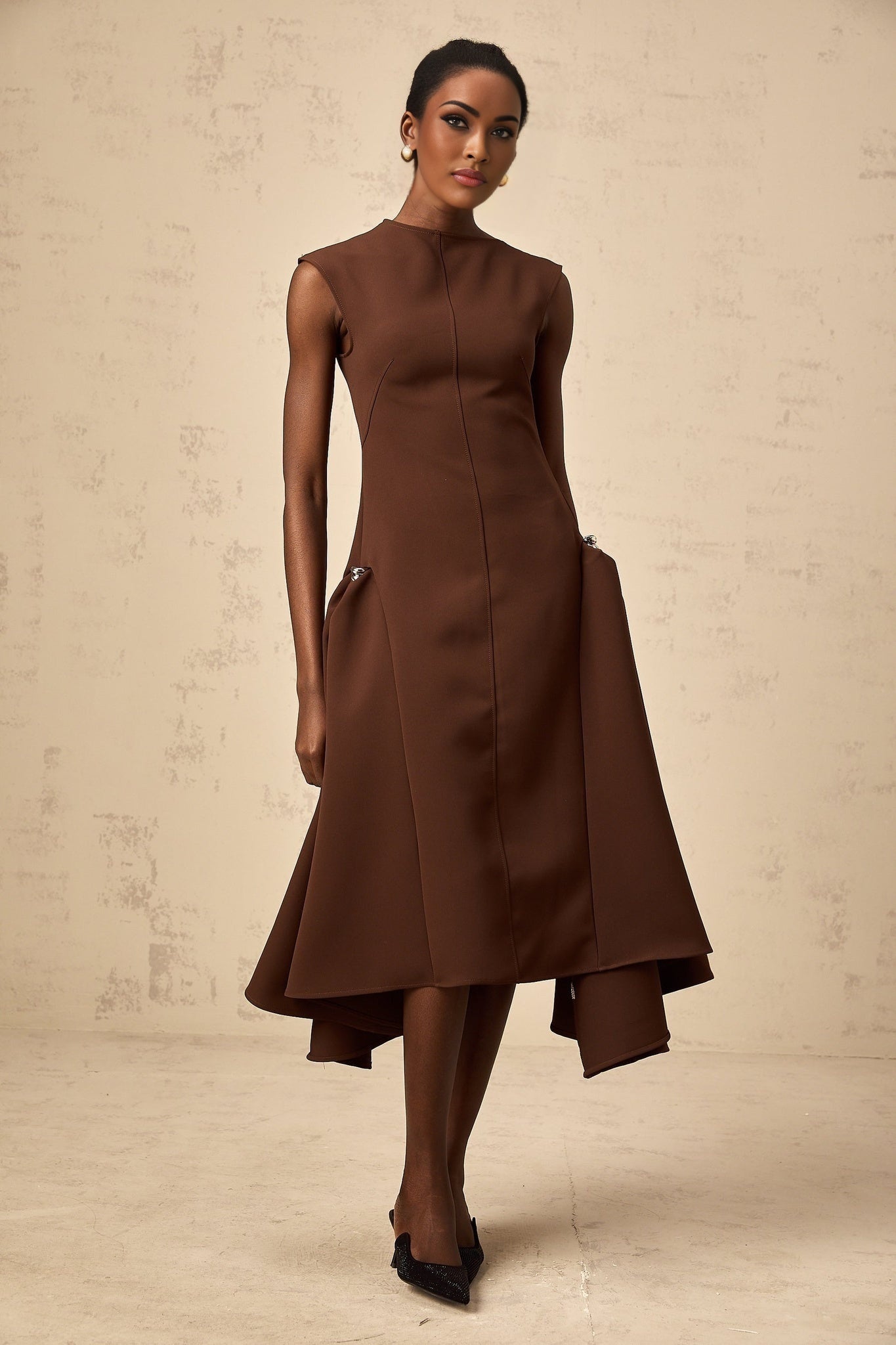 A woman wearing a brown asymmetrical midi dress with a flowing skirt and fitted bodice standing against a plain background