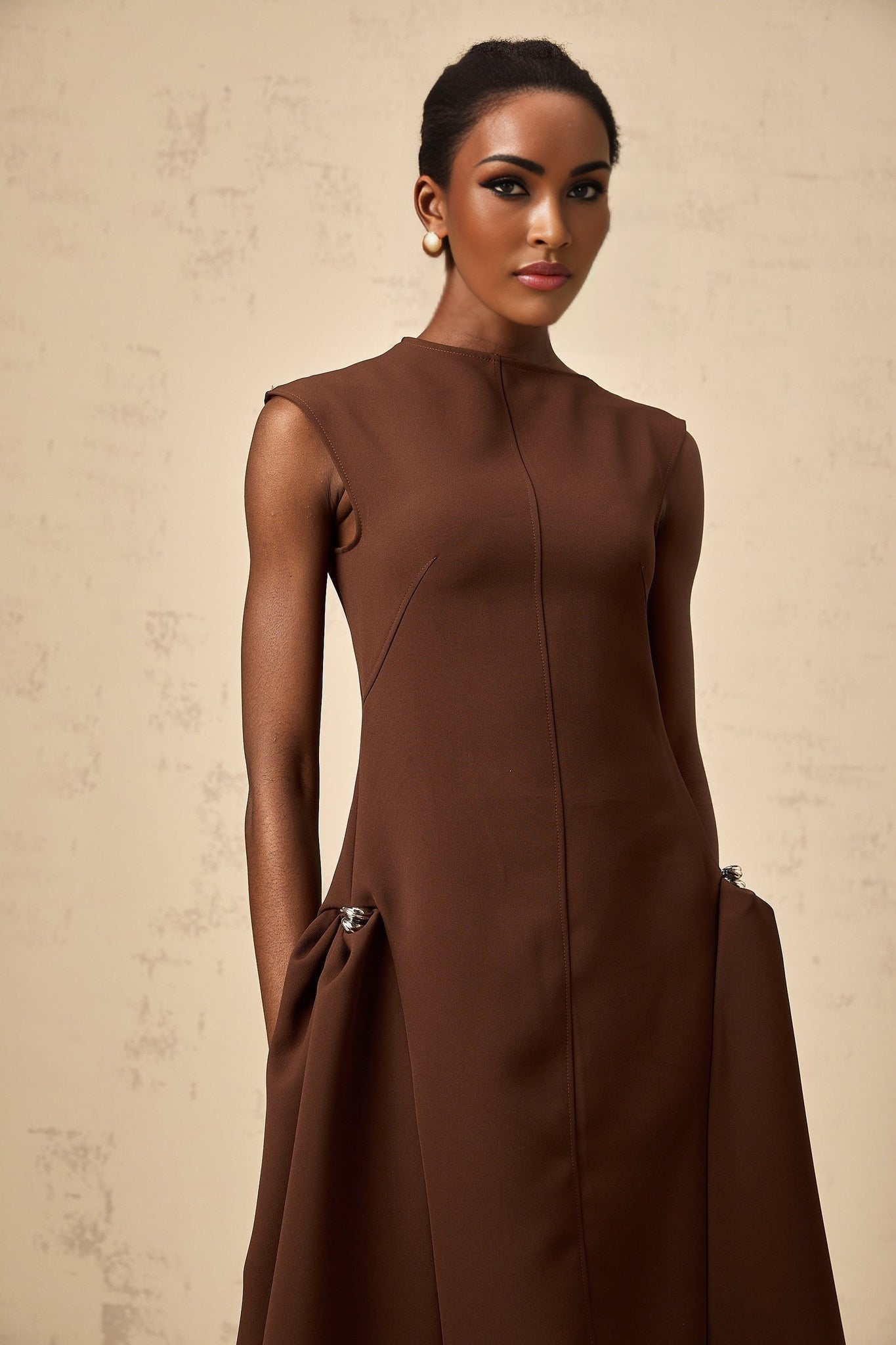 A woman wearing a brown asymmetrical midi dress with a flowing skirt and one shoulder design standing against a plain background