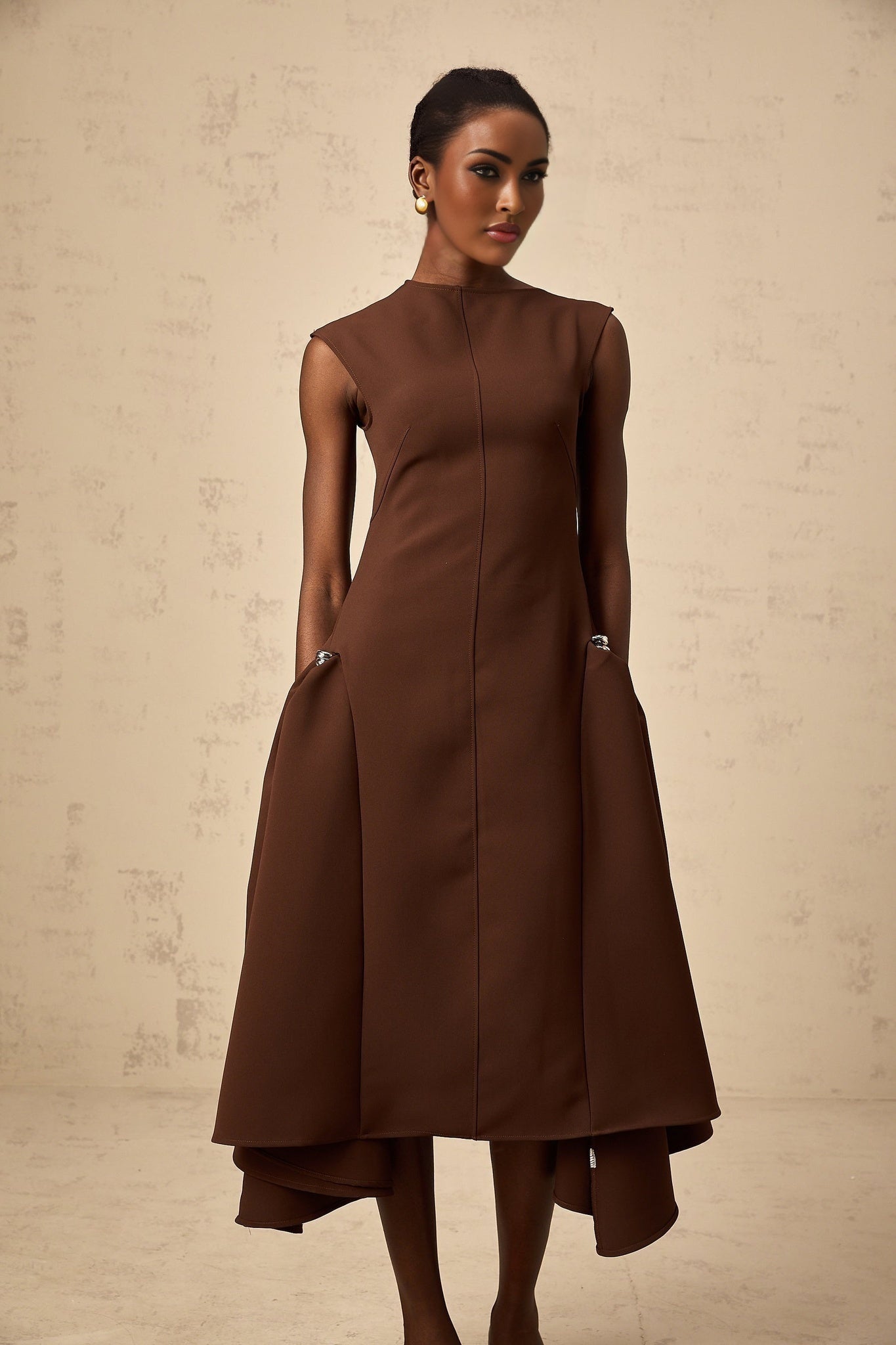 A woman wearing a brown asymmetrical midi dress with a flowing skirt and one shoulder design standing against a plain background