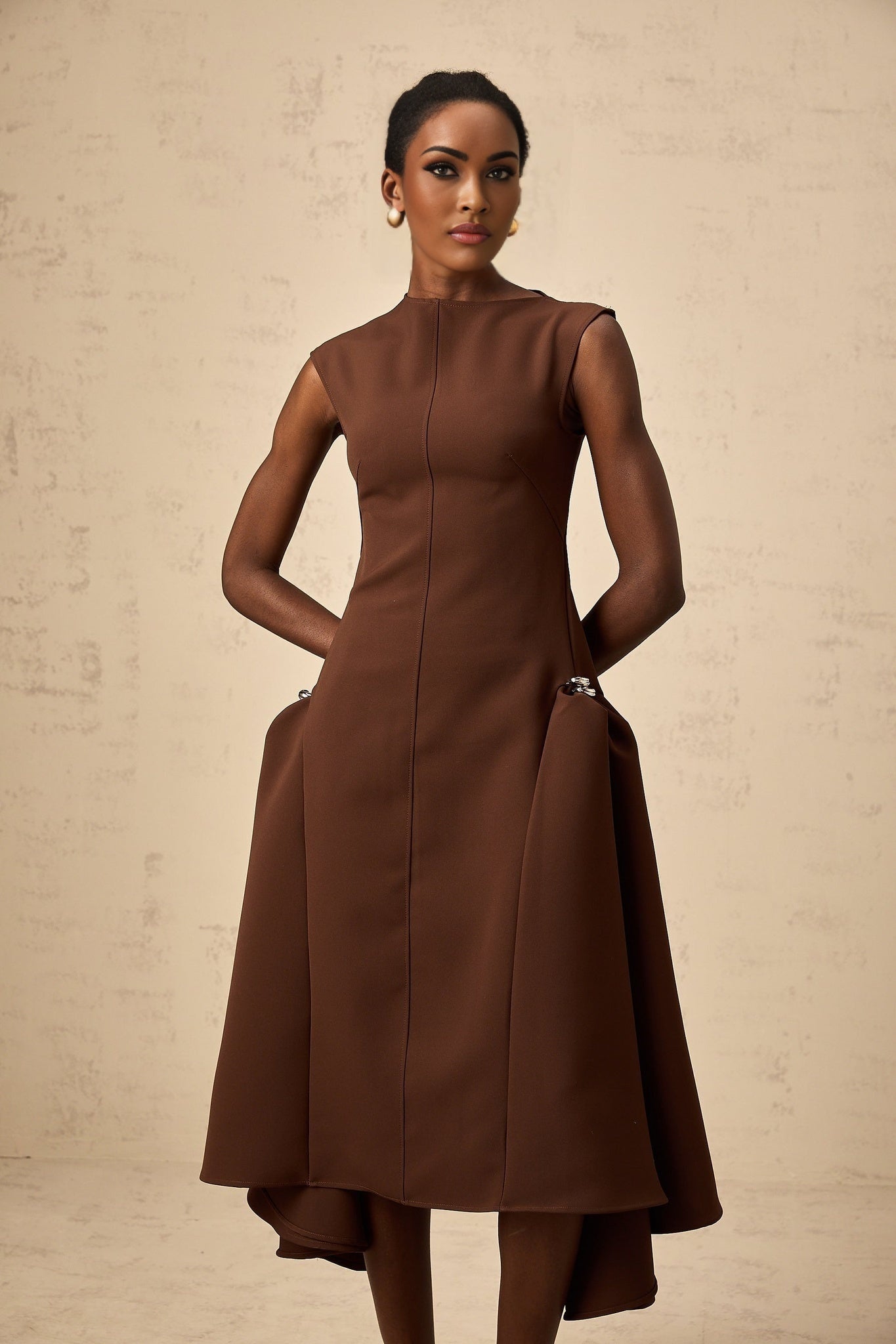 A woman wearing a brown asymmetrical midi dress with a flowing skirt and one shoulder design standing in a neutral pose