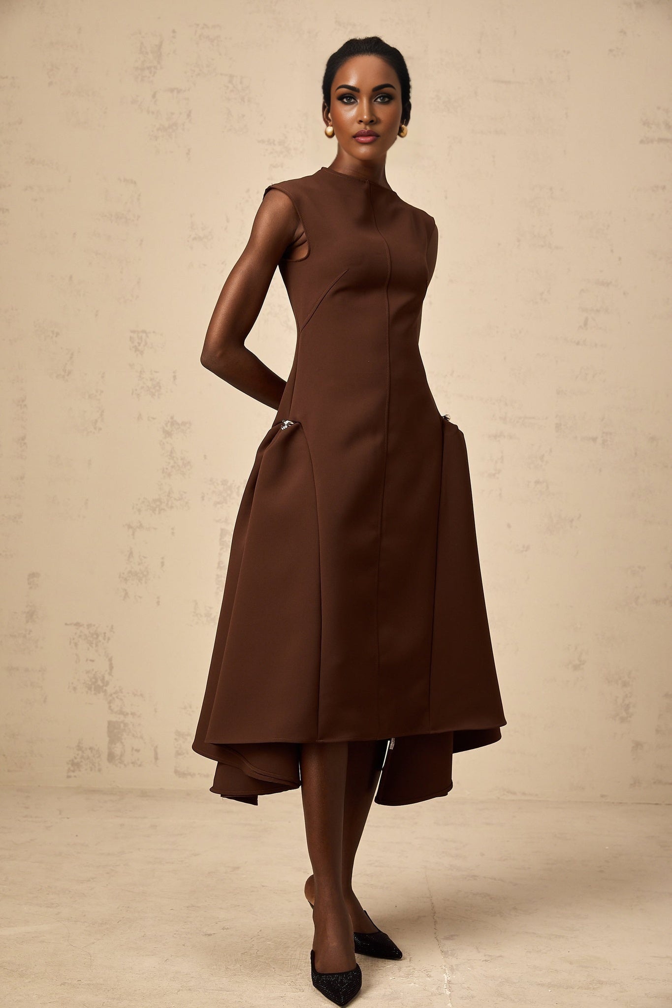 A woman wearing a brown asymmetrical midi dress with a flowy skirt and fitted bodice standing against a plain background