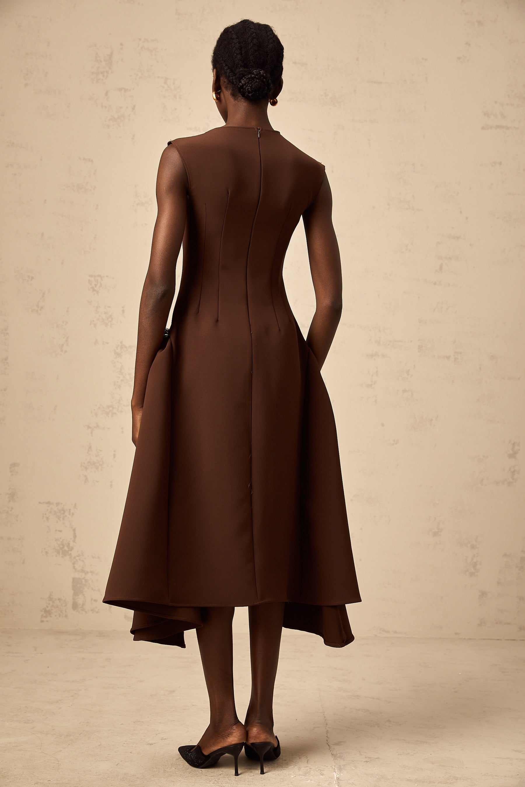 A woman wearing a brown asymmetrical midi dress with one shoulder exposed standing against a plain background