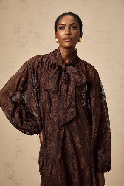 A woman wearing a brown flared midi dress made of chantilly lace named Sylvie The dress features a fitted bodice and a flowy skirt that falls just below the knee